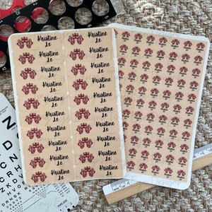 Pode incluir: Duas folhas de autocolantes com o nome "Kristine Le" e um gráfico de exame de vista. Os autocolantes são bege com texto e gráficos bordô. Ferramentas de exame de vista e uma régua também são visíveis.