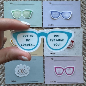 Może przedstawiać: Ręka trzymająca niebieską naklejkę z okularami z napisem "NOT TO BE CORNEA... BUT EYE LOVE YOU!" otoczoną innymi naklejkami i kartkami z okularami. Karty mają etykiety "TO" i "FROM".