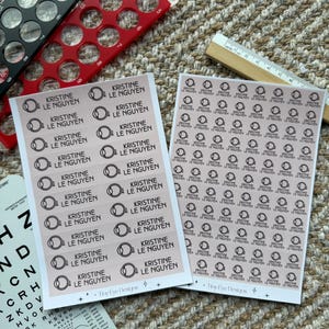 May include: Two sheets of light pink rectangular stickers featuring a black eye icon and the text "KRISTINE LE NGUYEN". The sheets are displayed alongside a black and red pupil gauge, a white eye chart, and a wooden ruler showing inches and centimeters.