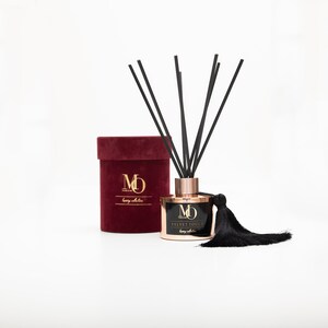 Puede incluir: Un frasco difusor negro y dorado con un borla negra y varillas difusoras negras. La botella está etiquetada con "Velvet Touch" y "MO Perfumes". El difusor está en una caja de terciopelo burdeos con el mismo logotipo.