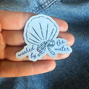 May include: A light blue sticker in the shape of a seashell with the words "washed by the water" in a handwritten font. The sticker has a holographic, glittery effect.