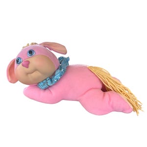 May include: A pink plush toy dog with blue eyes, a pink nose, and a blue patterned collar. The toy has a yellow yarn tail and ears. The dog is lying down on a white background.