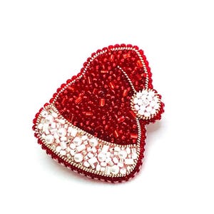 May include: A beaded Santa hat brooch in red, white, and gold. The hat is covered in red beads, with a white beaded trim and a white pom-pom. The brooch is likely handmade, ideal for festive occasions.