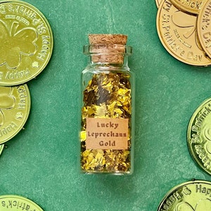 Op de afbeelding: Een klein glazen flesje gevuld met goudvlokken, met het label "Lucky Leprechaun Gold", staat op een groene ondergrond, omringd door gouden munten met klaverontwerpen en de tekst "Happy St. Patrick's Day".