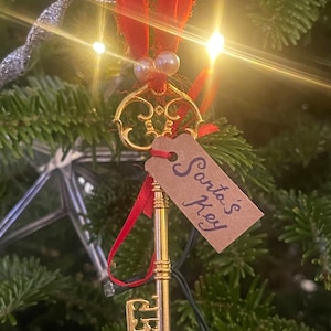May include: A gold-colored key ornament with a red ribbon and a brown tag that says "Santa's Key" hanging on a Christmas tree.
