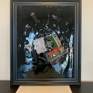May include: A black framed artwork featuring a clear glass bottle with a green label that reads "Salty Watermelon Whiskey" inside a black resin background.