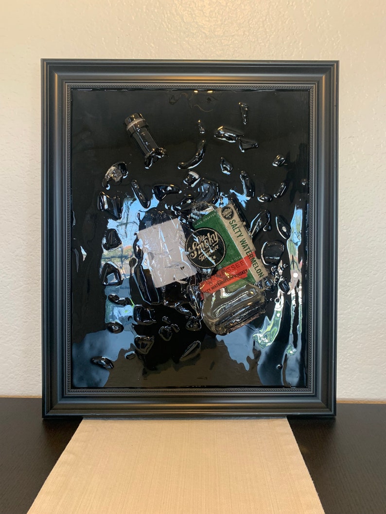 May include: A black framed artwork featuring a clear glass bottle with a green label that reads "Salty Watermelon Whiskey" inside a black resin background.