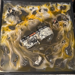 May include: A square resin art piece featuring a partially broken tequila bottle and fragments, with a label that reads "Espolon Tequila Blanco Mexico." The artwork incorporates swirling gold, white, and gray tones, creating a dynamic and abstract composition within a black frame.