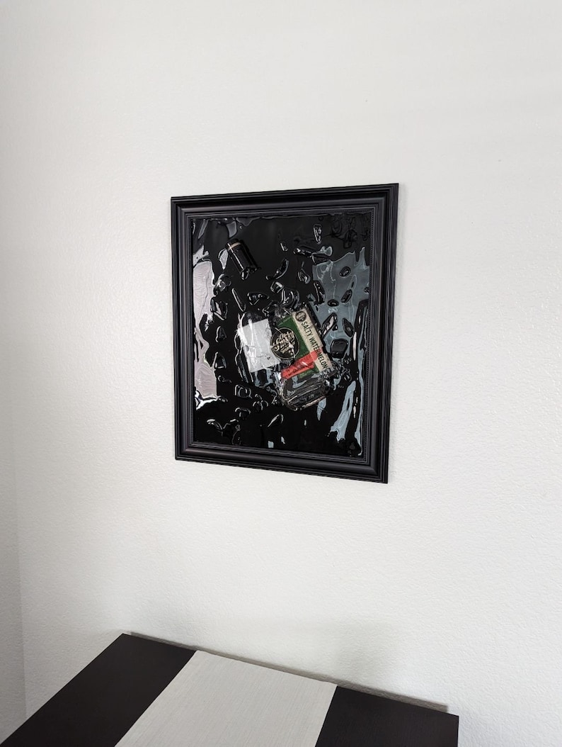 May include: A black framed picture with a clear resin background. A glass bottle and a small red, white, and green label are embedded in the resin. The label reads "SAFETY INSTRUCTIONS".