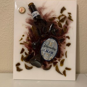 May include: A white canvas with a broken glass bottle of Yuengling Light Lager beer embedded in resin. The bottle is broken into many pieces, and the label is visible. The bottle is surrounded by a dark purple resin, and there are small pieces of glass scattered around the bottle.