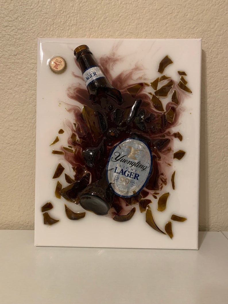 May include: A white canvas with a broken glass bottle of Yuengling Light Lager beer embedded in resin. The bottle is broken into many pieces, and the label is visible. The bottle is surrounded by a dark purple resin, and there are small pieces of glass scattered around the bottle.
