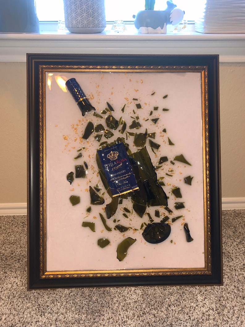 May include: A framed artwork featuring a broken glass bottle of Stella Rosa blueberry wine. The bottle is surrounded by shards of glass and gold leaf. The artwork is set against a white background.