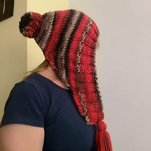 Op de afbeelding: Een handgebreide muts met een gestreept patroon in tinten rood, bruin en beige. De muts heeft een puntige vorm met een pompon aan de bovenkant en een lange, dikke kwast die aan de onderste punt hangt, gemaakt van grof garen.