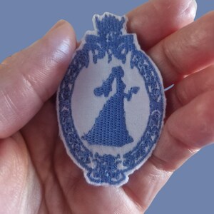 Puede incluir: Parche bordado azul y blanco con la silueta de una persona con un vestido largo sosteniendo un objeto. El parche tiene un borde decorativo y un diseño de corona en la parte superior. El parche se sostiene en una mano.