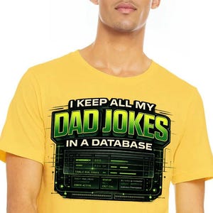 Op de afbeelding: Mosterdgeel T-shirt met de tekst "I KEEP ALL MY DAD JOKES IN A DATABASE" in groen en wit. Het ontwerp bevat een computerachtige afbeelding met groene accenten, wat een humoristische en technisch onderlegde uitstraling creëert.