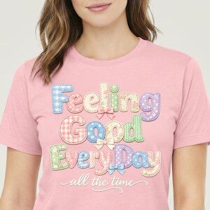 Può includere: T-shirt rosa chiaro con la scritta "Feeling Good Everyday all the time" in lettere colorate in stile patchwork. Le lettere sono in colori pastello con dettagli a fiocco.