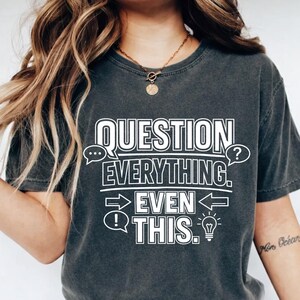 May include: Dark grey t-shirt featuring the white text "QUESTION EVERYTHING. EVEN THIS." The design includes speech bubbles, arrows, and a lightbulb. The shirt is a casual, comfortable style.
