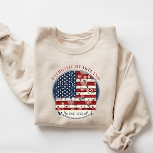 Op de afbeelding: Een crèmekleurige sweatshirt met een cirkelvormig patriottisch ontwerp. Het ontwerp bevat een blauwe cirkel met witte sterren, rood-witte strepen en de tekst "PATRIOTIC HEARTLAND EST. 1776".