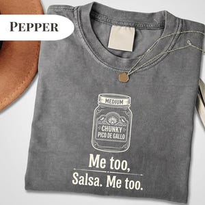 May include: A gray t-shirt with a white graphic of a jar labeled "Chunky Pico De Gallo" and the text "Me too, Salsa. Me too." A silver necklace with a small pendant is on the shirt.
