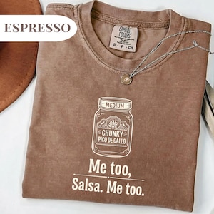 May include: Espresso-colored t-shirt with a crew neck and short sleeves. The shirt features a white graphic of a jar labeled "Chunky Pico de Gallo" and the text "Me too, Salsa. Me too." A silver necklace with a pendant is also visible.