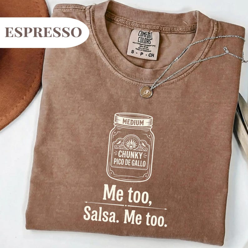 May include: Espresso-colored t-shirt with a crew neck and short sleeves. The shirt features a white graphic of a jar labeled "Chunky Pico de Gallo" and the text "Me too, Salsa. Me too." A silver necklace with a pendant is also visible.