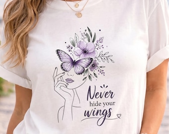May include: A white t-shirt featuring a black line art illustration of a face with a hand resting near the chin. A large purple butterfly covers the eye area, surrounded by purple flowers and leaves. The text reads Never hide your wings.