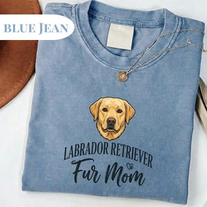 May include: Blue jean colored t-shirt featuring a Labrador Retriever graphic. The shirt has the text "LABRADOR RETRIEVER Fur Mom" in black script. A gold necklace with a pendant is draped on the shirt.