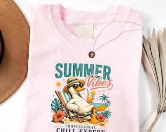 May include: A light pink t-shirt featuring a graphic of a duck wearing sunglasses and a straw hat, lounging in a beach chair with a drink. The text reads SUMMER VIBES and PROFESSIONAL CHILL EXPERT, surrounded by palm trees, flowers, and a sun.