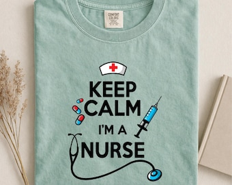 May include: A light sage green t-shirt featuring a graphic with the text KEEP CALM I'M A NURSE in black letters. The design includes a white nurse cap with a red cross, three small red and blue capsules, a blue syringe, and a black stethoscope.