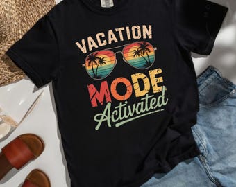 May include: A black t-shirt featuring a graphic of aviator sunglasses with a sunset and palm tree design inside the lenses. The text VACATION MODE Activated is printed in a retro font above and below the sunglasses in shades of orange and green.