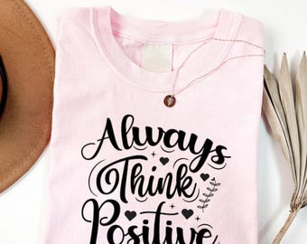 May include: A light pink crew neck t-shirt featuring the black text Always Think Positive in a decorative script font. The design includes small heart icons, star accents, and a vertical leaf sprig. A delicate gold-tone necklace rests on the shirt.