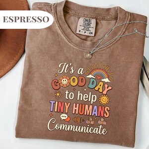 Puede incluir: Camiseta color espresso con el texto "It's a GOOD DAY to help TINY HUMANS Communicate" en una fuente retro. La camiseta tiene cuello redondo y mangas cortas. Se ve un collar plateado con un colgante.