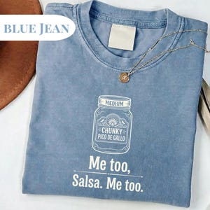 May include: Blue jean colored t-shirt with a white graphic of a jar labeled "Chunky Pico De Gallo" and the text "Me too, Salsa. Me too." A gold necklace with a pendant is draped across the shirt.