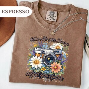 May include: Espresso-colored t-shirt with a floral camera graphic. The graphic includes a vintage camera surrounded by daisies, lavender, and other flowers. The text reads "When life gets blurry adjust your focus."
