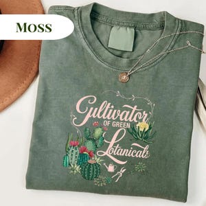 Op de afbeelding: Een mosgroen t-shirt met de tekst "Cultivator of Green Botanicals" in roze script, met een botanische illustratie van cactussen en bloemen. Een zilveren ketting met een hanger is over het shirt gedrapeerd.