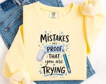 May include: A pale yellow crewneck sweatshirt featuring a graphic print with the text "MISTAKES ARE PROOF THAT YOU ARE TRYING" in black and blue, accompanied by illustrations of a grey eraser and a blue pencil with a silver ferrule.