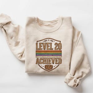 May include: A cream-coloured sweatshirt with a graphic that reads "LEVEL 20 ACHIEVED." The graphic features a shield design with a rainbow stripe and a trophy with the number 20. The sweatshirt has long sleeves and a crew neck.