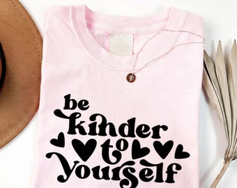 May include: A light pink t-shirt featuring the black text 'be kinder to yourself' in a stylized, rounded font. The design includes several black heart icons interspersed among the words. A delicate gold-toned necklace rests on the neckline.
