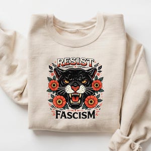 May include: Beige sweatshirt featuring a black panther graphic with the words "RESIST FASCISM". The panther is surrounded by red and orange flowers. The design has a vintage, tattoo-inspired aesthetic.