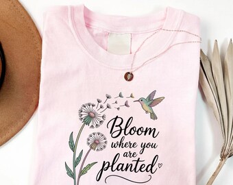 May include: A light pink t-shirt featuring a graphic of two dandelions with seeds blowing in the wind, a small hummingbird, and the black cursive text "Bloom where you are planted" with a small heart icon. The word "BLOSSOM" is printed in the top left corner.