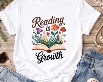 May include: A white short-sleeved t-shirt featuring a graphic of an open book with colorful flowers, green leaves, and mushrooms growing out of the pages. The text Reading is Growth is printed in a dark brown script font above and below the graphic.