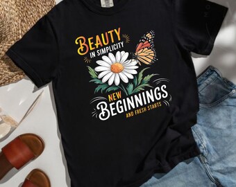 May include: A black short-sleeve t-shirt featuring a graphic print of a white daisy flower and an orange monarch butterfly. The text reads Beauty in Simplicity, New Beginnings, and Fresh Starts in white and yellow lettering on the front.
