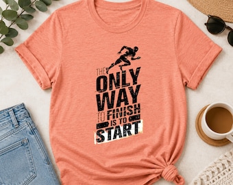 May include: A peach-colored short-sleeve t-shirt featuring a black graphic of a running figure and the text THE ONLY WAY TO FINISH IS TO START. The shirt has a crew neckline and a standard fit, showcasing a distressed, textured print design.
