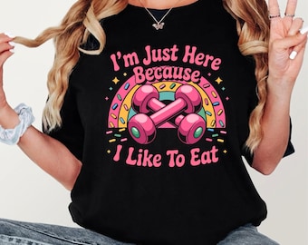 May include: A black t-shirt featuring a graphic print of two crossed pink dumbbells centered under a colorful rainbow arch with sprinkles. The text reads, I'm Just Here Because I Like To Eat, in pink lettering above and below the dumbbells.