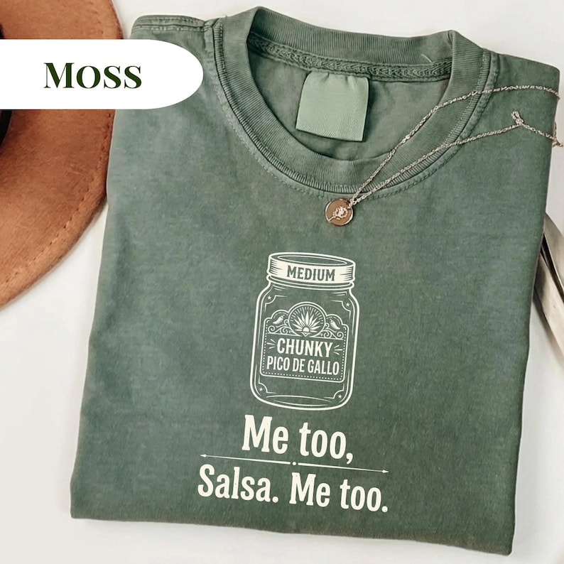 May include: Moss green t-shirt with a graphic of a jar labeled "Chunky Pico De Gallo" and the text "Me too, Salsa. Me too." A silver necklace with a small pendant is on the shirt.