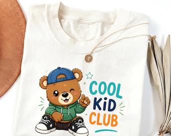 May include: An ivory t-shirt featuring a cartoon brown bear wearing a blue baseball cap, a green hoodie, and black and white sneakers. The text "COOL KiD CLUB" is printed in teal, dark blue, and orange next to the bear illustration.