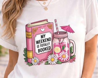May include: A white t-shirt featuring a graphic of a pink book and a pink mason jar drink with a straw and umbrella. The book has the text MY WEEKEND IS TOTALLY BOOKED. The design includes white daisies, green leaves, and yellow lemon slices.