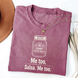 May include: A dusty rose-colored t-shirt with a crew neck. The shirt features a white graphic of a jar labeled "Chunky Pico De Gallo" and the text "Me too, Salsa. Me too." A gold necklace with a pendant is draped on the shirt.