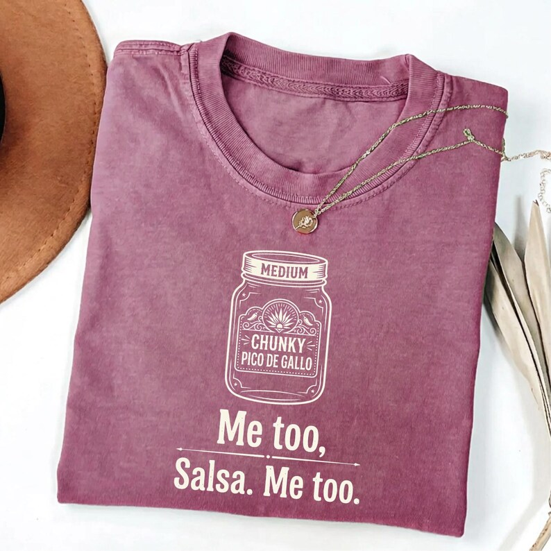May include: A dusty rose-colored t-shirt with a crew neck. The shirt features a white graphic of a jar labeled "Chunky Pico De Gallo" and the text "Me too, Salsa. Me too." A gold necklace with a pendant is draped on the shirt.
