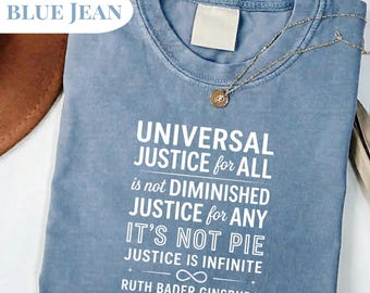 May include: A blue jean-colored t-shirt with the text "UNIVERSAL JUSTICE for ALL" and a quote by Ruth Bader Ginsburg. A gold necklace with a pendant rests on the shirt.